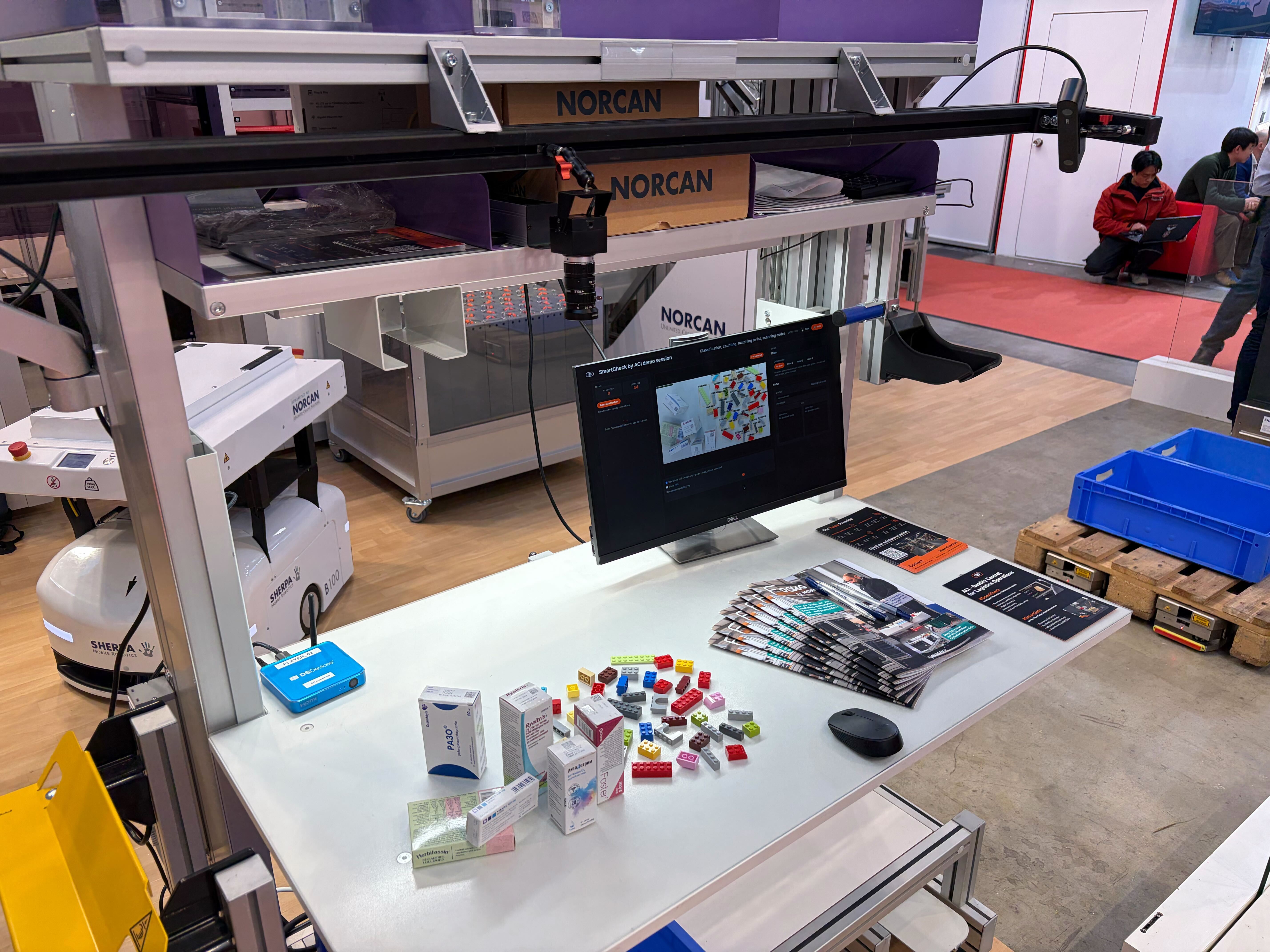 The ACI SmartCheck stand at LogiMAT 2026: an overhead camera on a frame above a white table, a monitor showing the interface, and products and parts laid out for the demo.